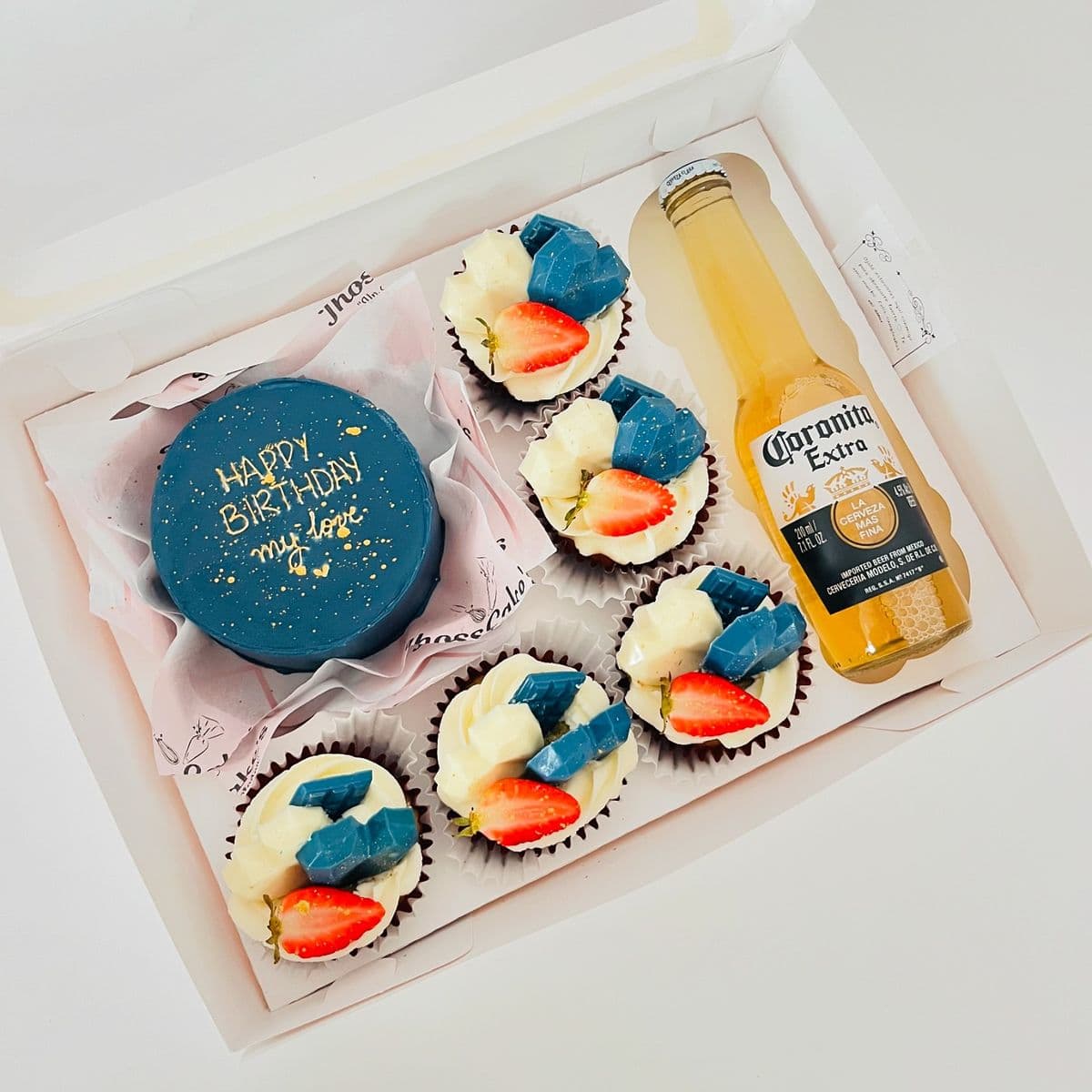Caja blanca con pastel azul de cumpleaños, cinco cupcakes decorados y una cerveza Coronita.