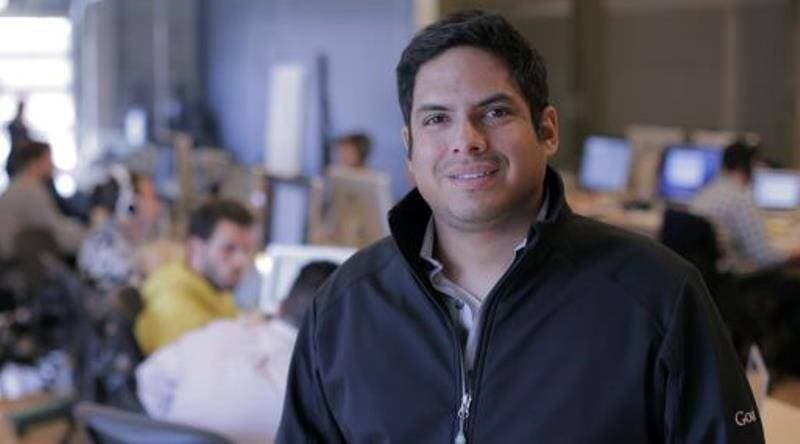 Hombre sonriente con chaqueta negra en una oficina moderna con colegas trabajando al fondo.