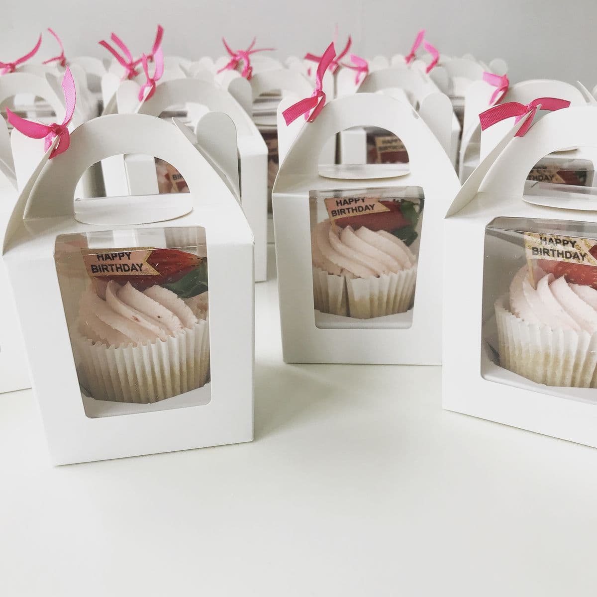 Cupcakes con glaseado rosa en cajas blancas individuales con lazos rosas y letreros de cumpleaños.