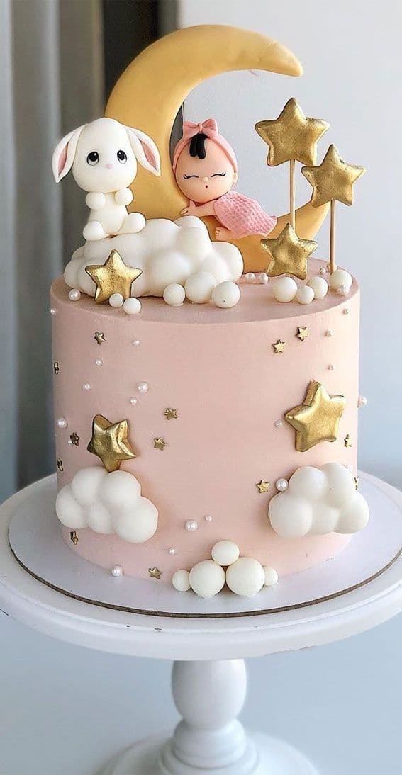 Pastel rosa decorado con luna, bebé durmiendo, conejo blanco, nubes blancas y estrellas doradas.