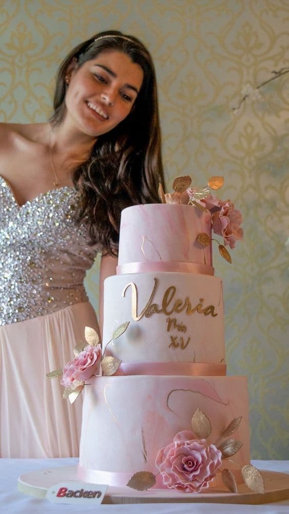 Quinceañera sonriente junto a un pastel rosa de tres niveles con flores y detalles dorados.