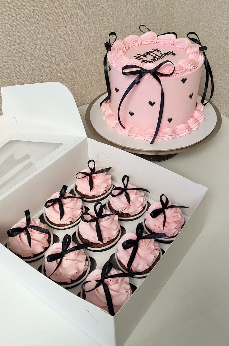 Pastel de cumpleaños rosa con lazos negros y corazones junto a una caja de cupcakes.