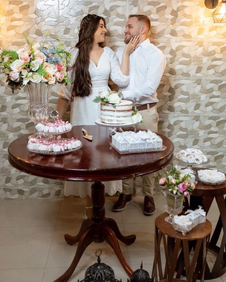Pareja vestida de blanco posando cariñosamente detrás de una mesa con pastel y flores.
